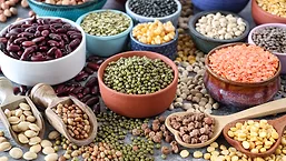 Close-up image group of dishes in a variety of sized bowls with pulses, nuts, seeds, peas and beans, black, green, red and yellow lentils, wooden serving scoops and heart-shaped spoons, spilled beans and lentils, grey background, focus on foreground
