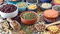 Close-up image group of dishes in a variety of sized bowls with pulses, nuts, seeds, peas and beans, black, green, red and yellow lentils, wooden serving scoops and heart-shaped spoons, spilled beans and lentils, grey background, focus on foreground