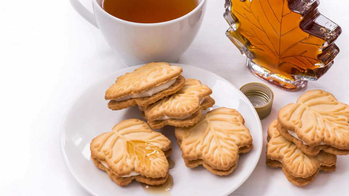 Maple leaf cookies with a creamy filling, maple syrup in a leaf-shaped bottle, and a cup of tea on a white tablecloth.