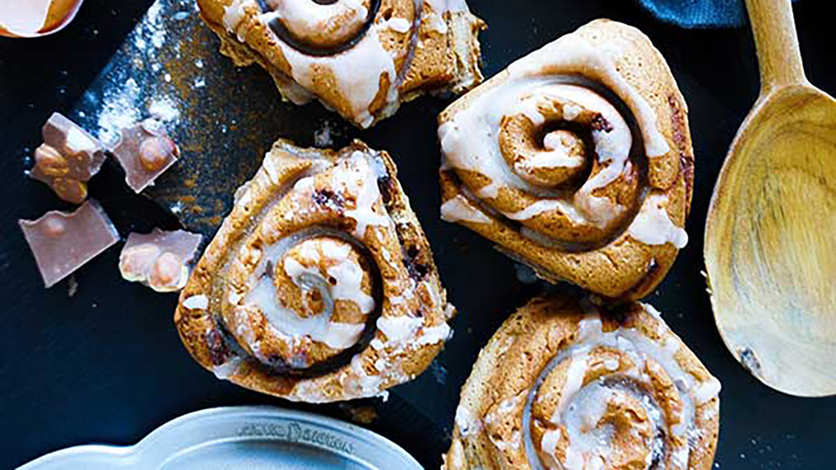 Four iced cinnamon rolls on a dark surface with eggshells, chocolate pieces, and a wooden spoon.