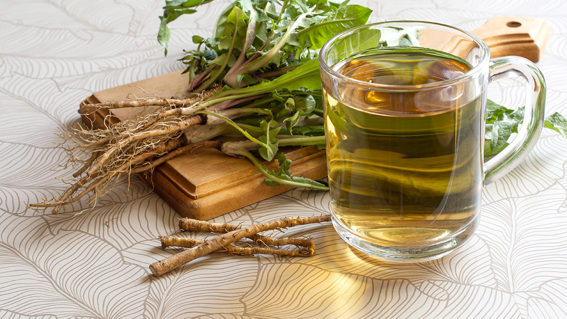 Fresh dandelion roots and leaves next to a clear mug of dandelion tea on a patterned surface.