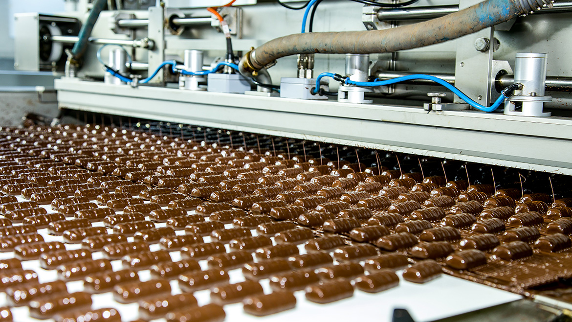 Rows of chocolate candies moving on a conveyor belt, being coated by industrial machinery in a factory.