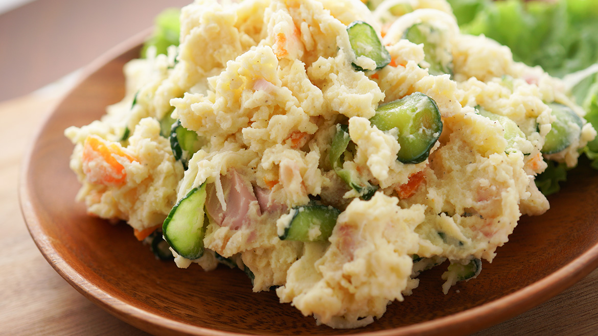 Creamy potato salad with cucumber, carrots, and ham on a wooden plate.