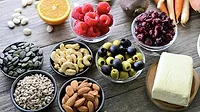 Small bowls of nuts, seeds, green and black olives, raspberries and cranberries, cream cheese, a half slice of an orange, more seach and various vegetables on the table.