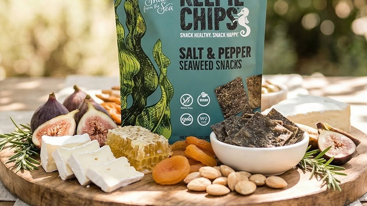 A bag of Snacks from the Sea Salt & Pepper Seaweed Snacks Kettle Chips on a wooden board, surrounded by figs, cheese slices, a block of honey, dried apricots, seeds and herbs.