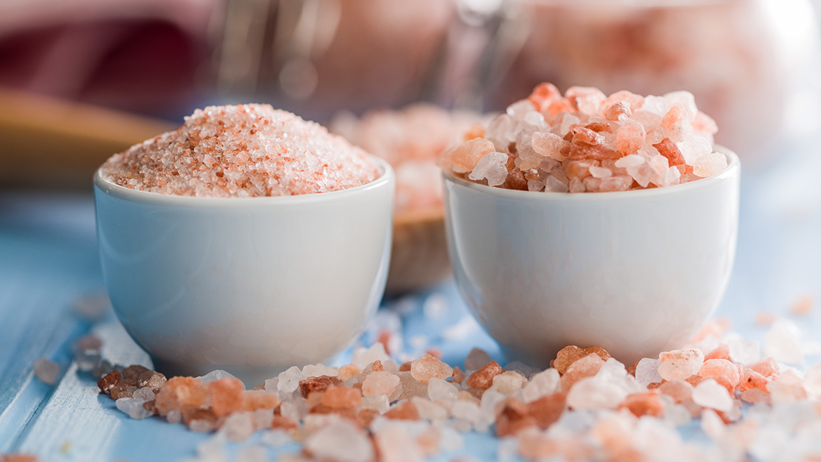 Two small white bowls of pink sea salt, one finely ground and the other in larger pieces. A wooden spoon is included in the blurred background.