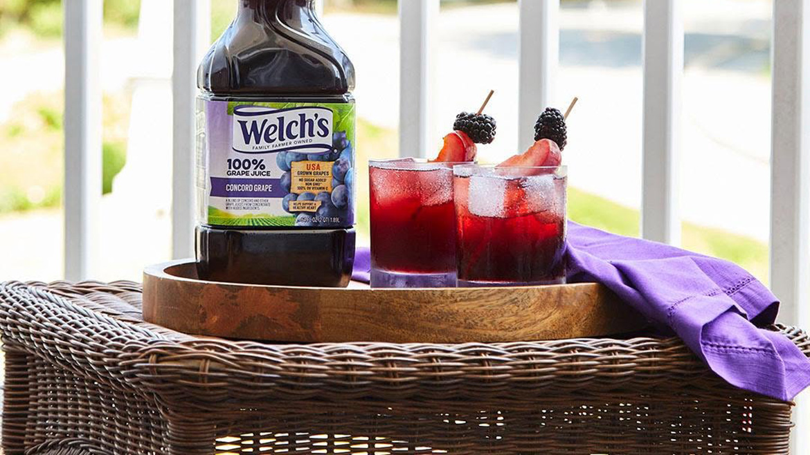 A bottle of Welch's 100% Grape Juice on a wooden tray along with two full glasses with ice and fruit garnish. A purple napkin lies on the tray. The tray is sitting on a rattan side table with a porch railing in the background showing the outdoors.