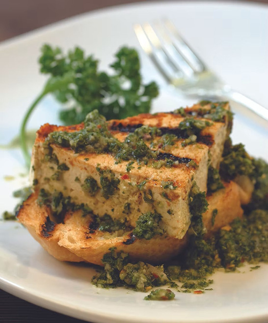 Grilled slices of tofu topped with an herb filling on a white plate with a garnish and a fork.