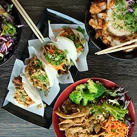 Overhead view of Asian-inspired rice bowls, bao buns, shrimp, chicken, fresh vegetables, and chopsticks arranged on a dark wooden table.