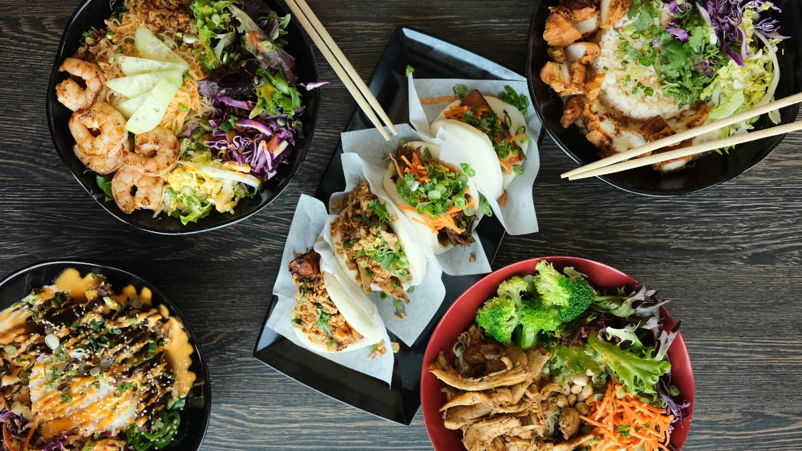 Overhead view of Asian-inspired rice bowls, bao buns, shrimp, chicken, fresh vegetables, and chopsticks arranged on a dark wooden table.