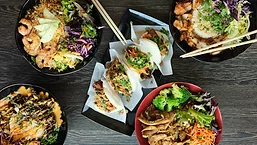 Overhead view of Asian-inspired rice bowls, bao buns, shrimp, chicken, fresh vegetables, and chopsticks arranged on a dark wooden table.